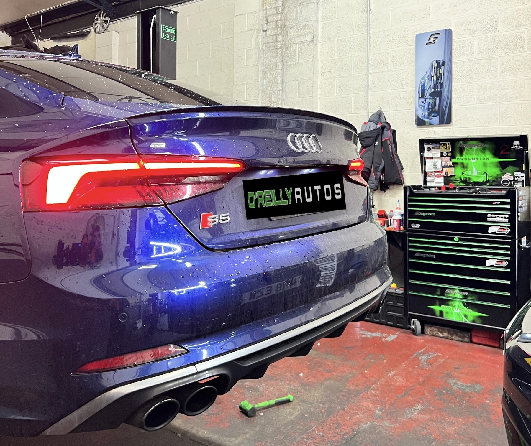 The back of a dark blue Audi S5 in the workshop wearing an O'Reilly Autos plate, with green tool chests behind