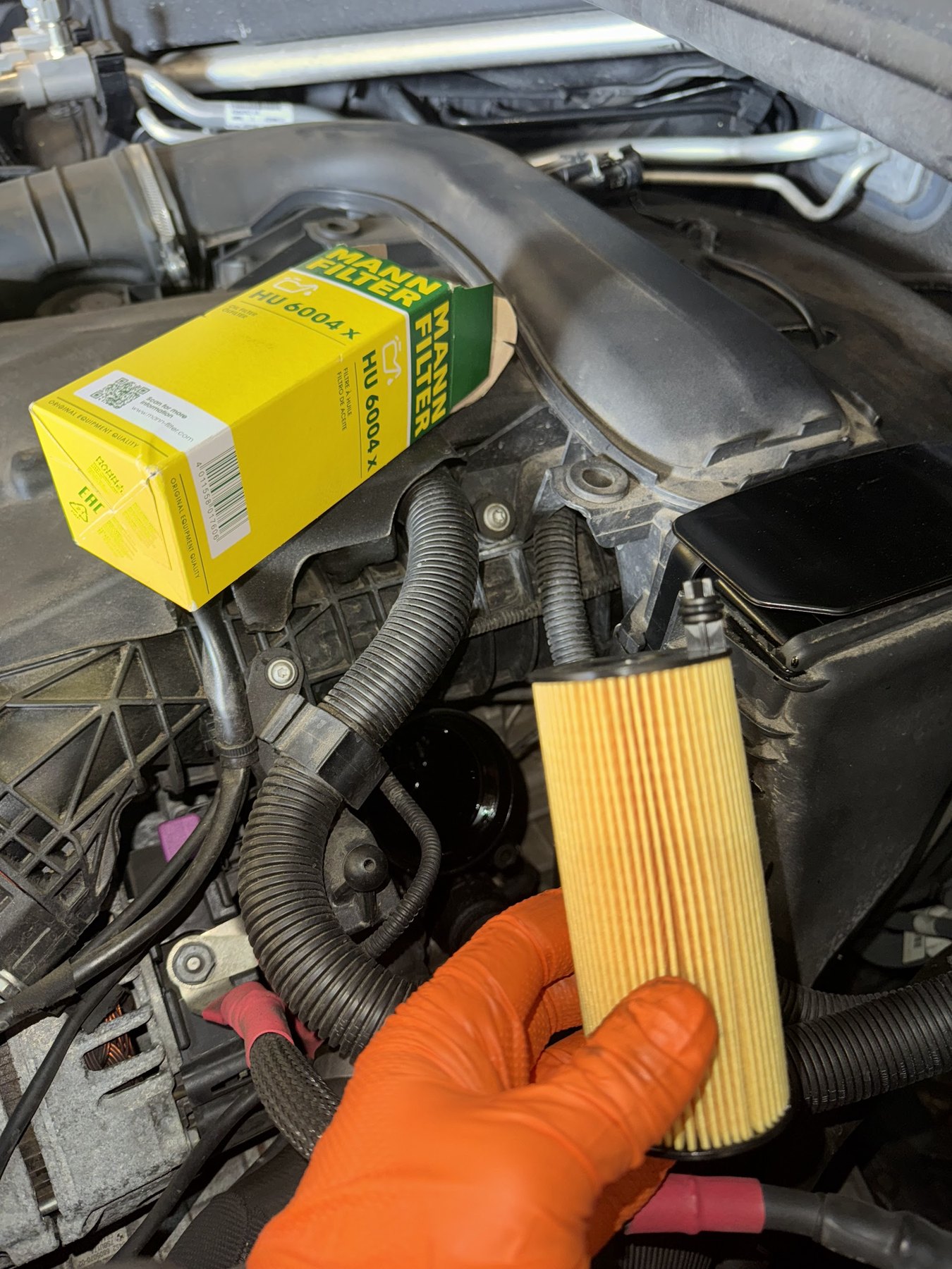 A used oil filter element held in an orange glove beside its boxed replacement, over an engine