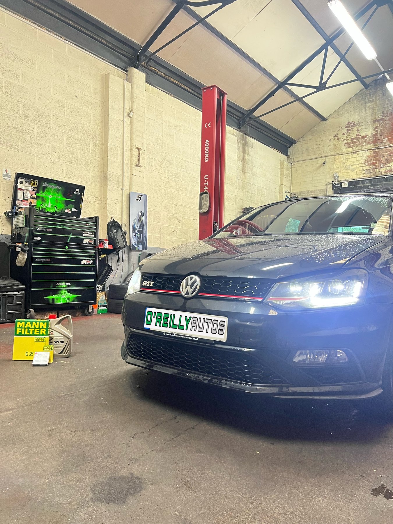 A Volkswagen Polo GTI in the workshop with its lights on, a green tool chest behind